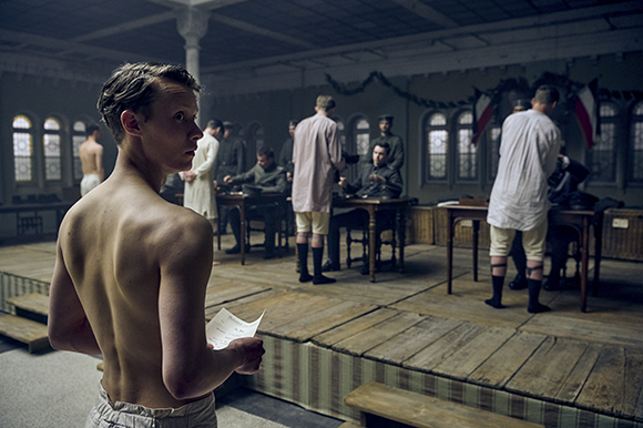A shirtless man holds a paper in a large, dimly lit room where several men sit at desks.