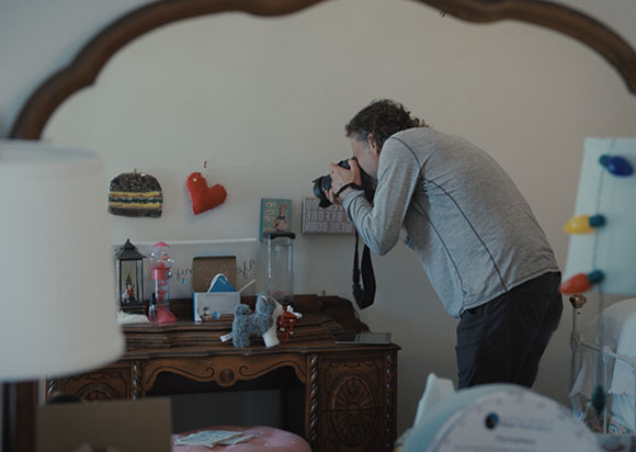 A man in a grey long-sleeve shirt leans forward to take a photo of objects on a wooden desk.