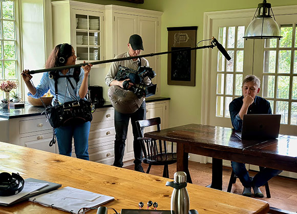 A woman holds a boom pole while a man operates a camera and a man sits at a table with a laptop.