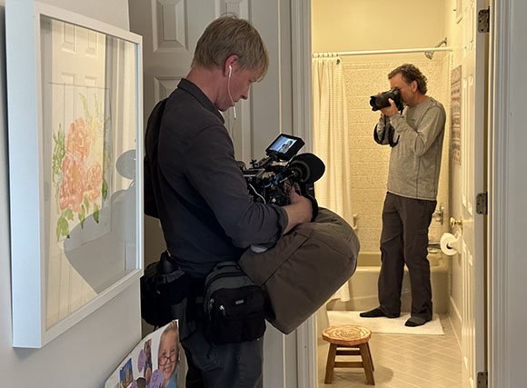 A man holds a video camera in a hallway while another man takes a photo in a bathroom through an open door.