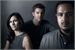 A woman and two men pose for a moody, low-light portrait against a dark grey background.
