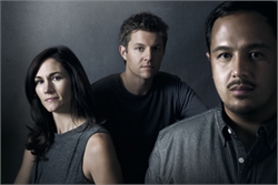 A woman, a man in a black shirt, and a man in a grey shirt looking at the camera against a dark backdrop.