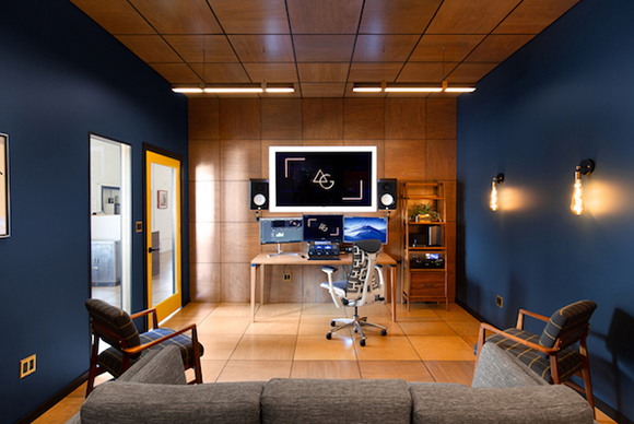 A media editing studio with a wooden desk, multiple monitors, studio speakers, and blue walls.