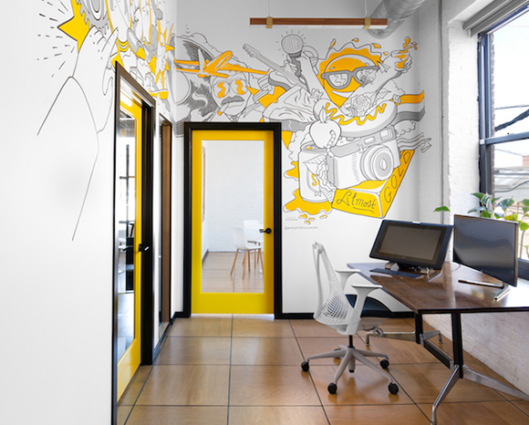 Office interior with a yellow door frame, a desk with monitors, and a large yellow and black line-art mural.