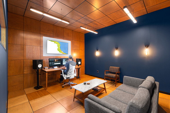 A person sits at a wooden desk with dual monitors in a studio with wood paneling and a blue accent wall.