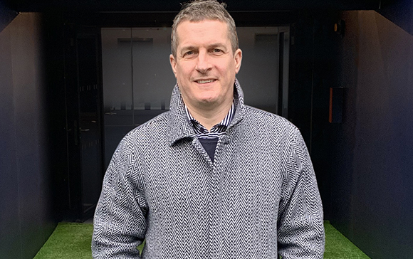 A man smiling while wearing a grey herringbone patterned jacket over a striped shirt.