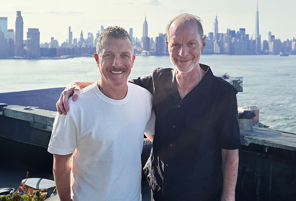 Two men smiling with their arms around each other on a rooftop overlooking a city skyline.