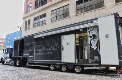 A large black mobile laboratory trailer with Alpha Lab branding parked next to a brick building.