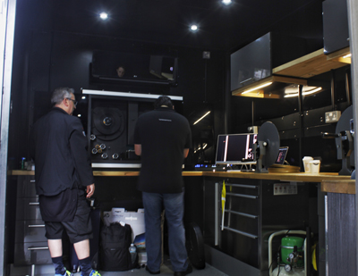 Two men in black clothing work inside a dark, high-tech mobile production trailer with monitors and equipment.