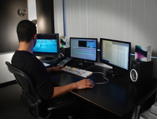 A person sits at a dark desk using three computer monitors, a keyboard, and speakers.