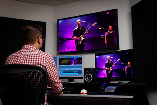 A person in a red checkered shirt monitors video footage on three screens in a dark studio.