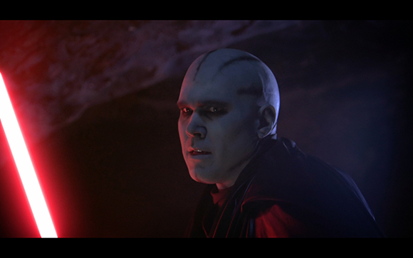 A pale man with facial markings looks intensely toward a glowing red lightsaber in a dark setting.