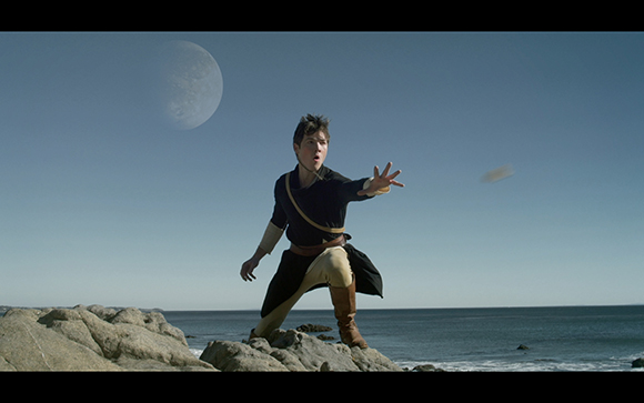 A man in black and tan clothing poses on coastal rocks under a clear sky with a large moon visible.