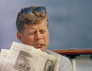 A man with sunglasses on his head reads a newspaper and smokes a cigar outdoors.
