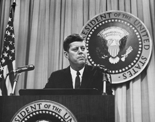 John F. Kennedy stands behind a podium with the Seal of the President of the United States in the background.