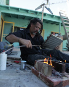 A man in a black shirt sits outdoors near a small fire built between bricks, holding a metal component.
