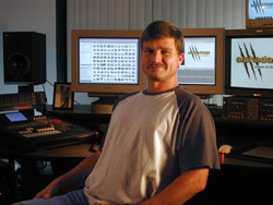A man with a mustache sits in a studio surrounded by computer monitors and audio mixing equipment.