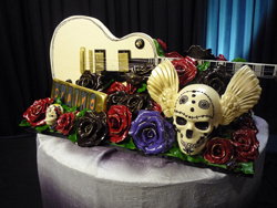 A guitar-shaped cake decorated with red, purple, and chocolate roses and a large winged sugar skull.