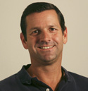 A smiling man with short dark hair wearing a dark blue polo shirt against a plain background.
