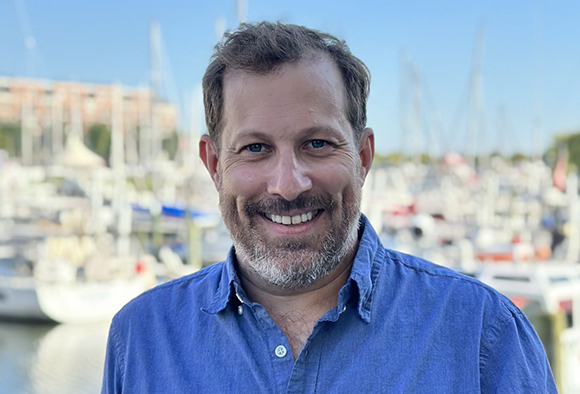 A smiling man with short brown hair and a salt-and-pepper beard wearing a blue button-down shirt.