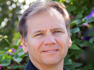 A man with light brown hair and blue eyes smiling against a background of green leaves and purple flowers.