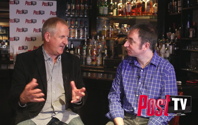 Two men sit at a bar, one gesturing while speaking to the other.