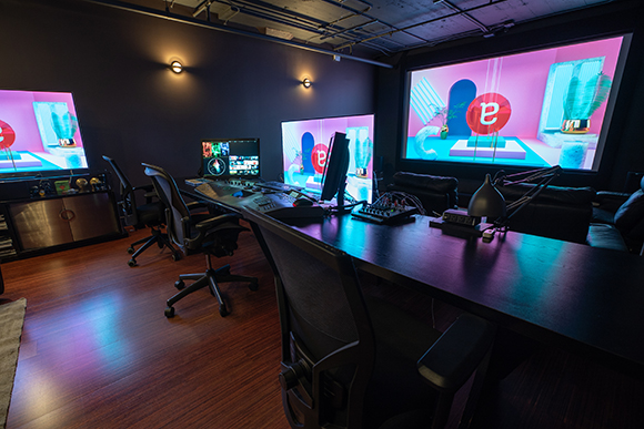 A dark video editing studio featuring multiple large screens displaying colorful graphics and a long black desk.
