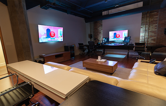 A lounge area with a white sofa and table next to a production studio with monitors and desks.