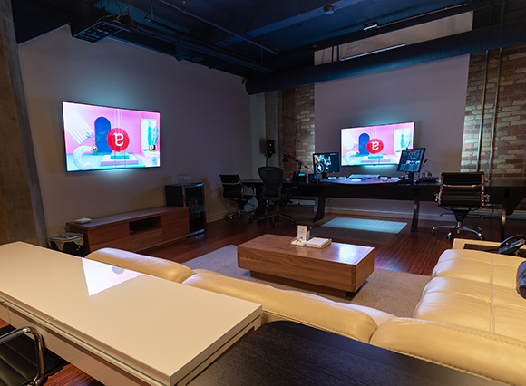 A dark production studio featuring large wall-mounted screens, workstations, and a white leather sofa.