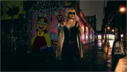 A woman in a black corset and sunglasses stands on a wet city street next to colorful graffiti.