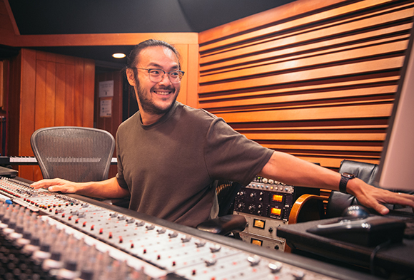 A smiling man with glasses and long hair operates a large audio mixing console in a wood-paneled studio.