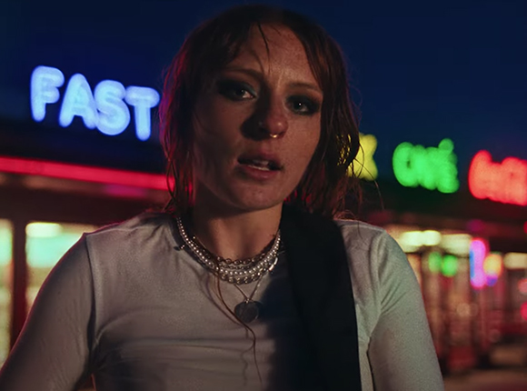 A woman with reddish hair and a white top stands in front of blurred neon lights at night.