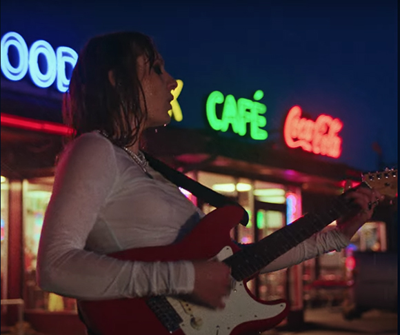 A person in a white long-sleeve shirt plays a red electric guitar in front of glowing neon cafe signs.