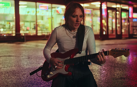 A woman with reddish hair plays a red electric guitar on a street at night.
