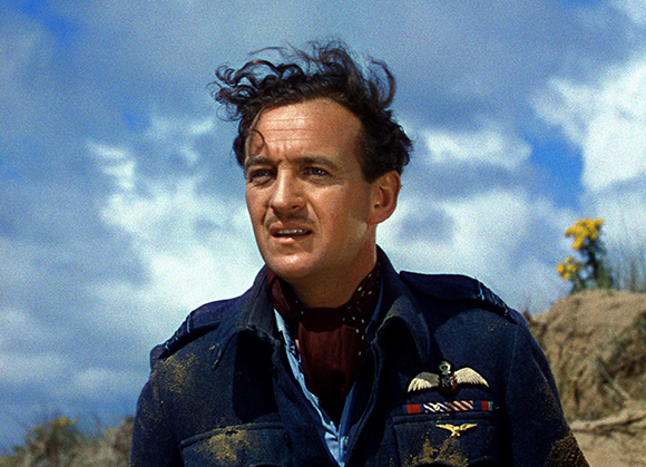A man with dark, wavy hair wearing a dark blue military uniform and a red neckerchief under a blue sky.
