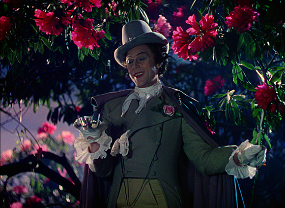 A person in a grey top hat and period clothing smiles while standing among blooming pink flowers.