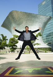 A man in a dark suit jumps with arms outstretched in front of a large, silver, sculptural building.