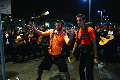 Two men in orange shirts dance joyfully in front of a large group of musicians playing guitars at night.