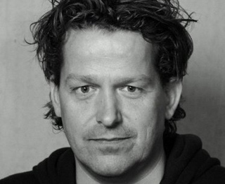 Black and white close-up portrait of a man with wavy hair looking directly at the camera.