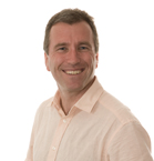 A smiling man with short brown hair wearing a light peach button-down shirt against a white background.