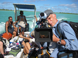 A man holding a large video camera with a monitor on a boat in turquoise water.