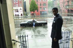 A man in a black suit stands on a balcony overlooking a wet street with a black limousine parked below.