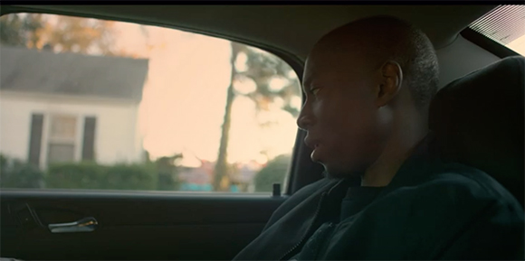 A man in profile sits in the backseat of a car, looking out the window at a house and trees.