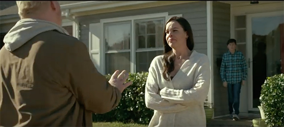 A man gestures toward a woman with crossed arms outside a house as a boy stands in the doorway.