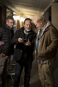 Three men stand in a dimly lit hallway, looking down at a smartphone held by the man in the center.