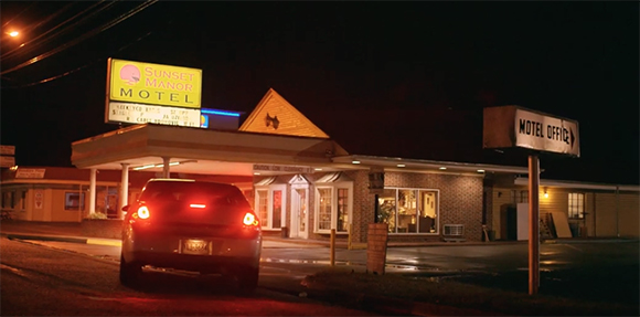 A car with glowing red taillights parked in front of the Sunset Manor Motel at night.
