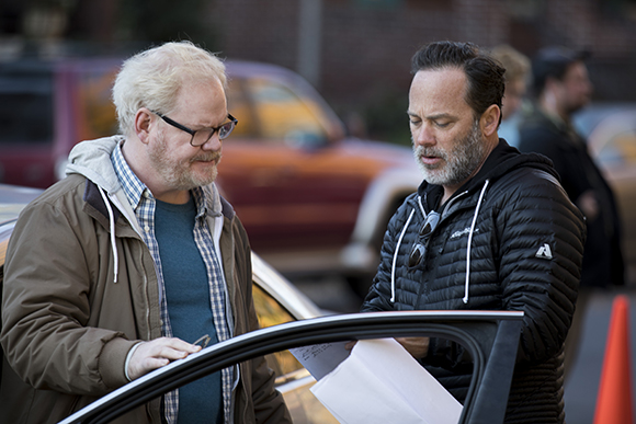 Two men stand near a car, one in a brown jacket and glasses, the other in a black puffer jacket, looking at papers.