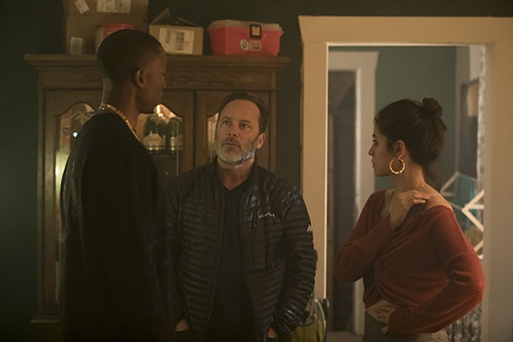 A man in a black jacket, a man in a dark shirt, and a woman in a red top talk in a room.