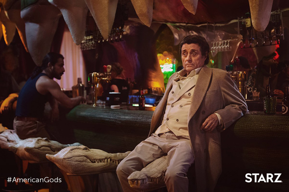 A man in a tan suit sits at a bar next to crocodile-shaped stools in a dimly lit room.