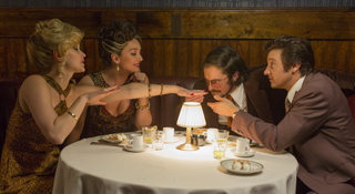 Four people in vintage attire lean toward a small, glowing lamp on a round table in a dark room.